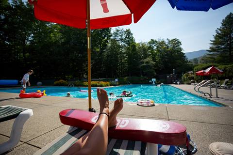 Legs on a lounge chair sitting pool side with umbrellas above 
