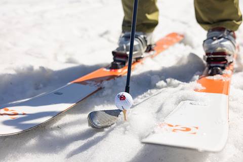 Feet on snow in ski boots on skis with a golf ball and club inbetween