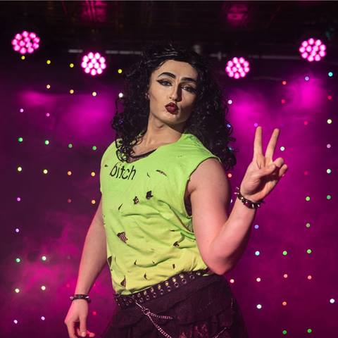 headshot of drag queen jizzo giving a peace sign in front of a pink light backdrop