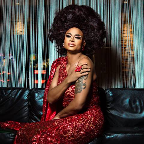 headshot of drag queen rhedd rhum in a long red dress in front of a blue tinsel backdrop