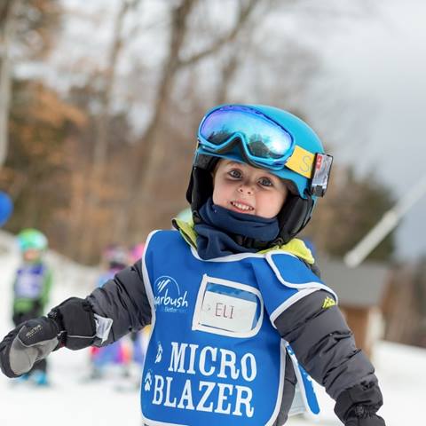 A little boy smiling at the camera wearing a helmet and bib that says micro blazer