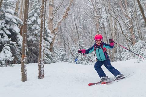A little girl in a multi colored jacket smiling and skiing in the woods