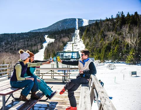 Group of people sitting on the Mount Ellen deck sharing a drink with a ski lift in the background