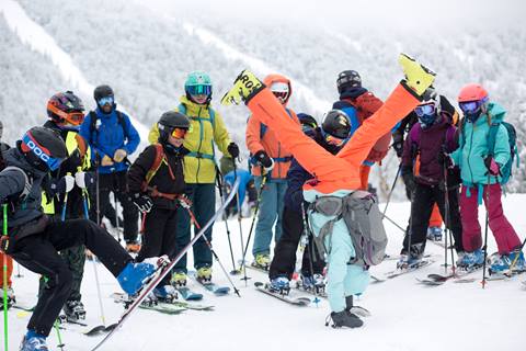 A ski group in a semi circle around a person doing a hand stand with ski boots on a snowy ski trail