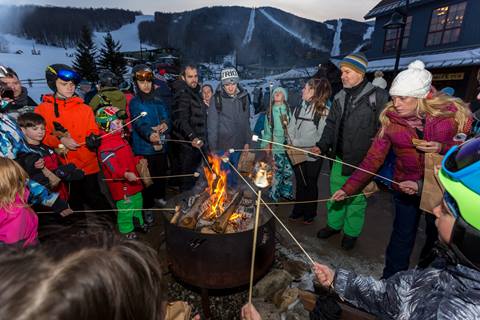 Group of people sanding around a fire pit toasting marshmellos outside of Gate House Lodge at Sugarbush Resort