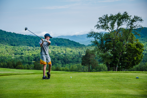 Junior golfer teeing off on a lush green course, wearing shorts and a short-sleeve shirt, mid-swing under clear skies.